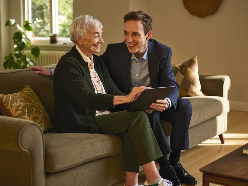 Illustration: Eine Seniorin und eine Begleiterin schauen gemeinsam auf ein Tablet im Wohnzimmer