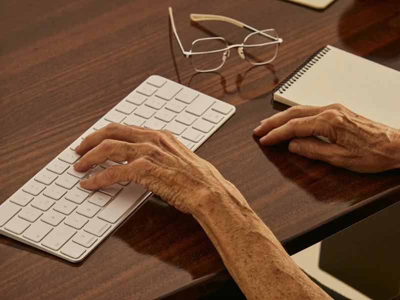 Illustration: Hände einer älteren Person ruhen auf einer großen Tastatur neben Lesebrille und Notizblock